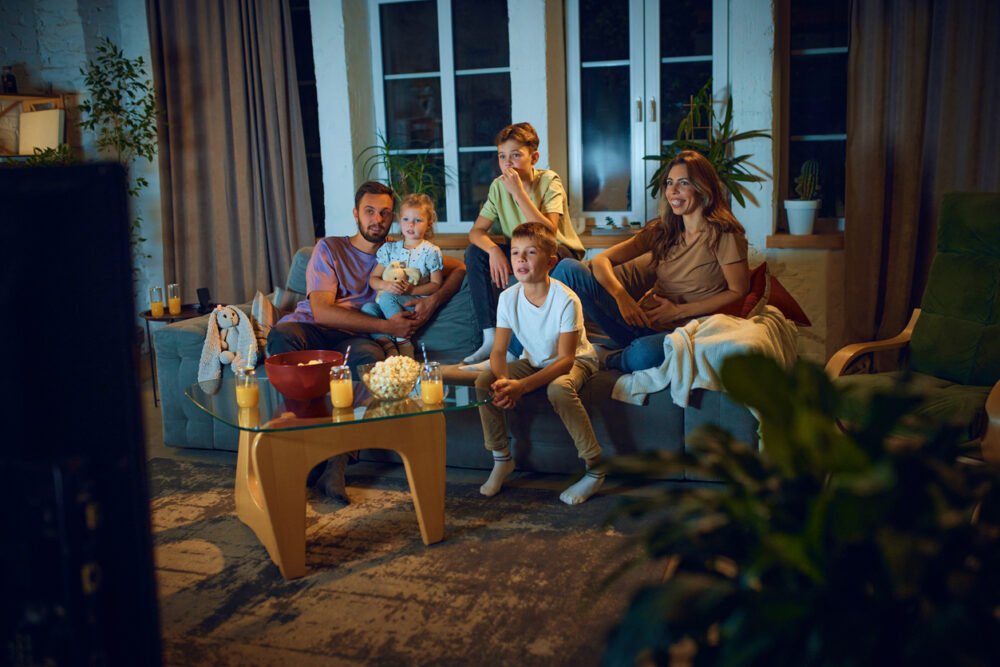 Living room, family watching TV, couch-centered scene, nighttime glow, documentary style, five people (three kids).