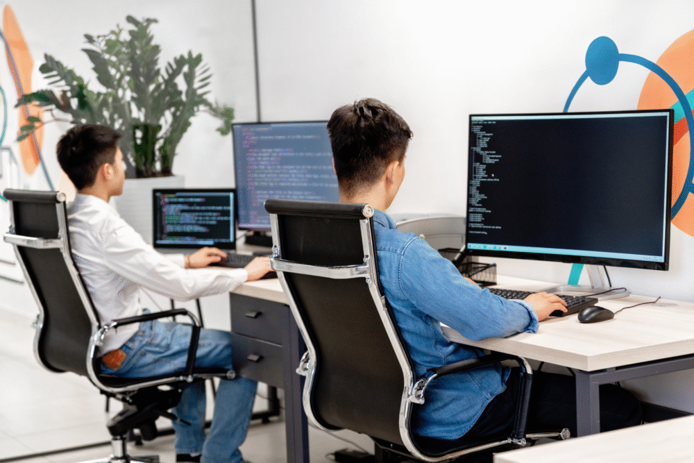 Modern office, dual desks, large code monitors, daytime natural light, editorial travel photo, two people present.