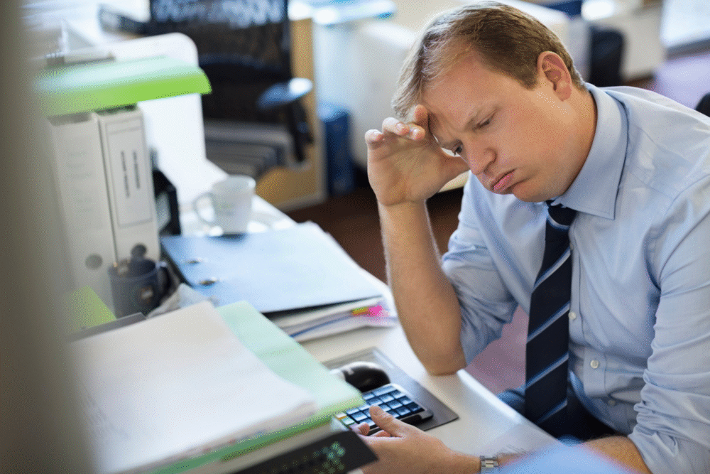 Office workspace, fatigued businessman, cluttered desk foreground, afternoon light, documentary style, one person.