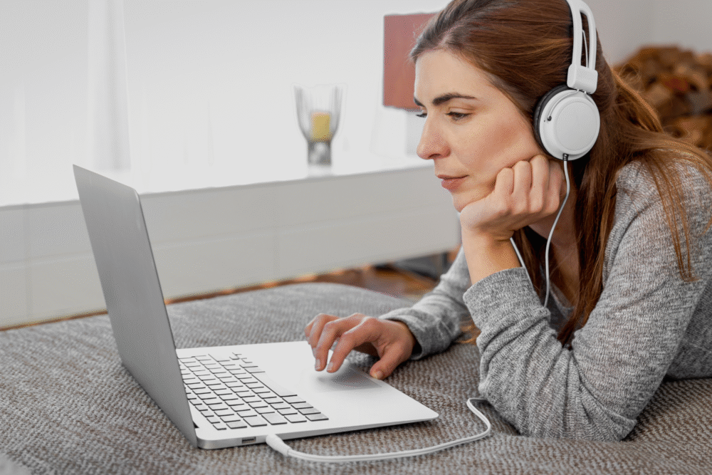 Bedroom, woman with headphones on bed using laptop, side angle, soft natural light, documentary style, one person.